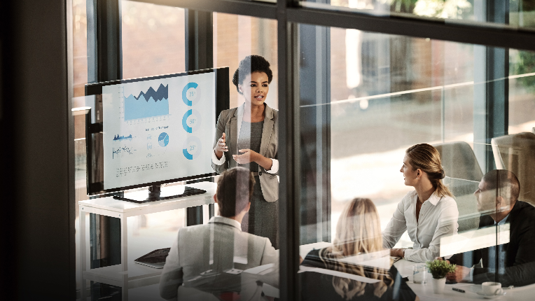 A lady standing beside a screen displaying various graphs presenting to people in a meeting room