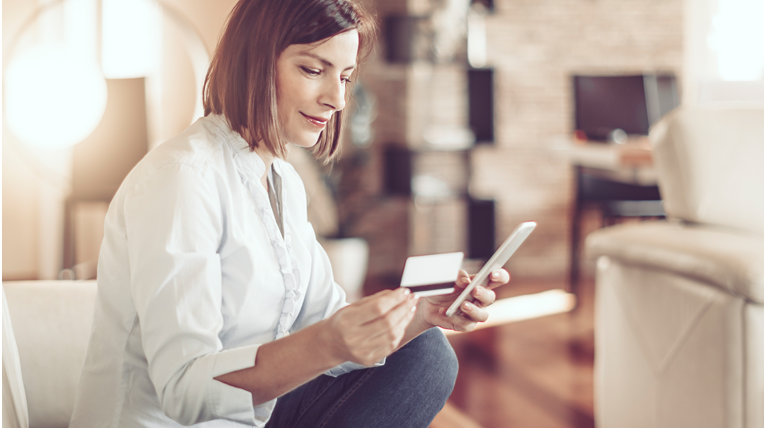 Assise sur un canapé blanc dans une salle de séjour, une femme tient une carte dans une main et un téléphone dans l’autre