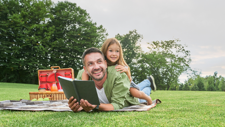Cheerful cute little girl spending time with her father, reading a book while having picnic in the park.