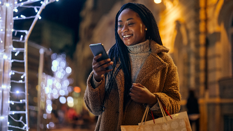 Jeune femme noire souriante se tenant dans les rues de la ville avec des sacs à provisions pendant la période de Noël et tapant sur son téléphone portable.