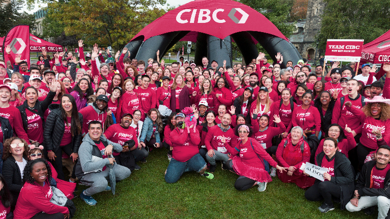 Crowd of people wearing red CIBC shirts outside on grass and man in the centre holding small pink dog to show support for raising money to fund cancer research.