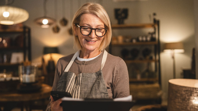 A person wearing an apron is standing indoors, holding a clipboard and a phone,