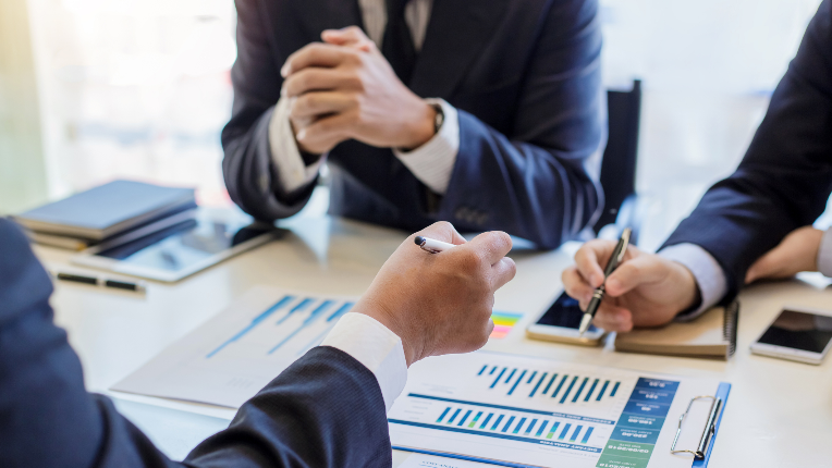 Three professionals sit amongst a table reviewing documents