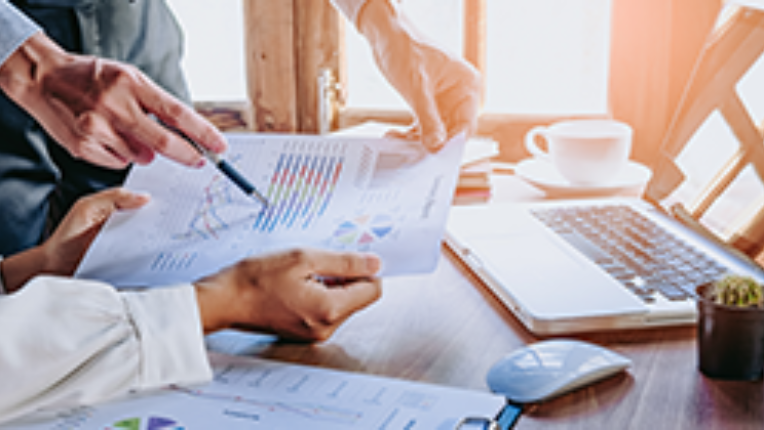 Two pairs of hands holding a paper with graphs over a desk with a laptop and cup of coffee