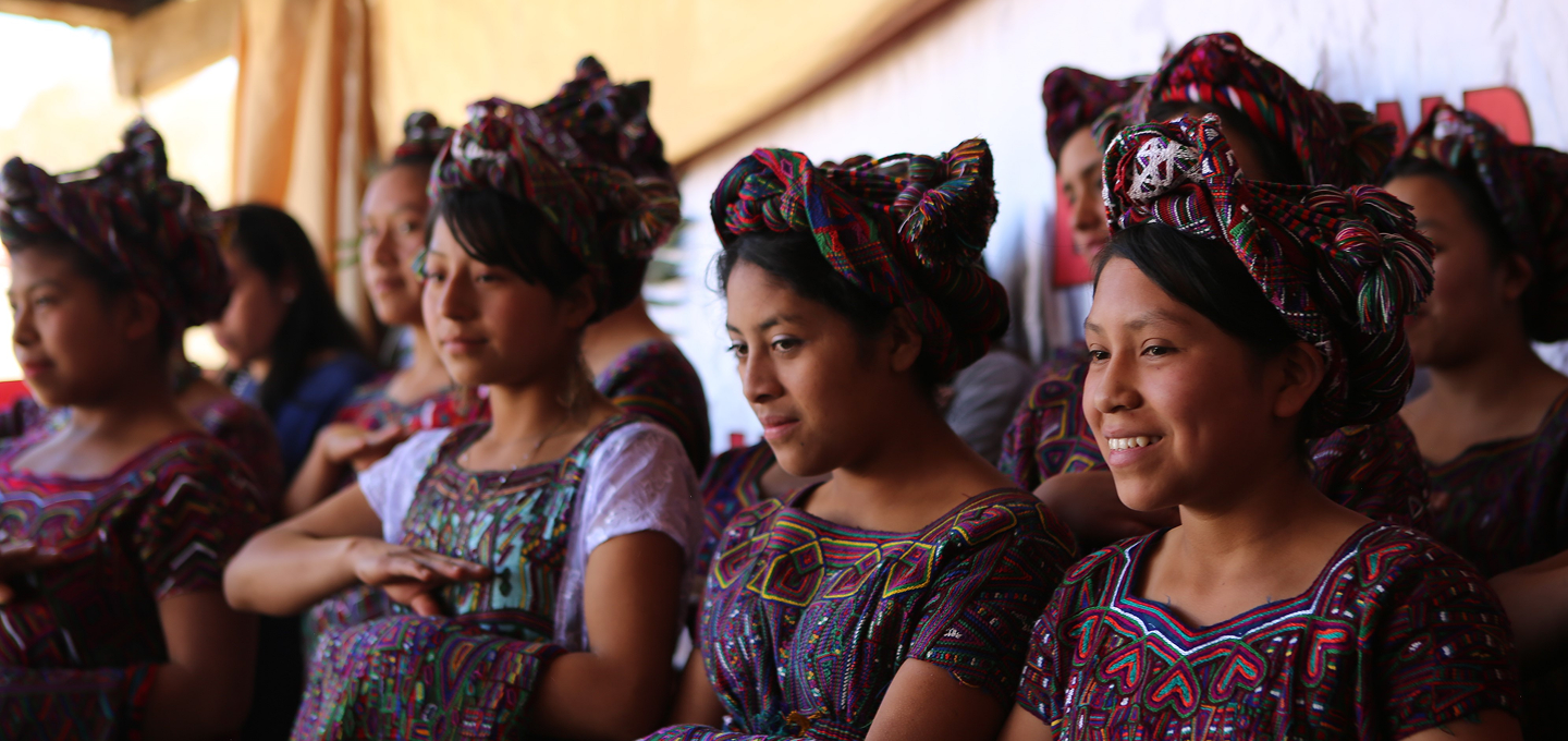 A group of individuals dressed in vibrant traditional clothing with intricate woven patterns in reds, purples, greens, and blues stand together indoors or under a partially covered area. Some wear matching headdresses or wraps tied in elaborate styles, and hold folded fabric pieces or shawls that match their outfits. Behind them is a white backdrop with partially visible red text and a large red decorative object resembling a paper fan or ribbon above the text. Light enters from the left side, and the overall mood is formal or ceremonial.