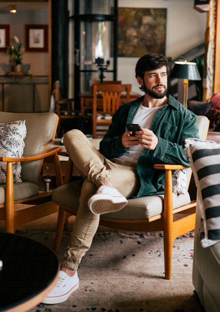 A young person sits casually in a modern lounge, holding a smartphone, surrounded by comfortable chairs and decor.
