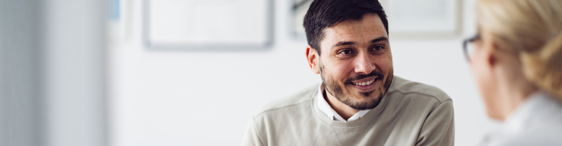 Smiling man in conversation indoors, facing a blurred person across from him in a bright office or meeting space.