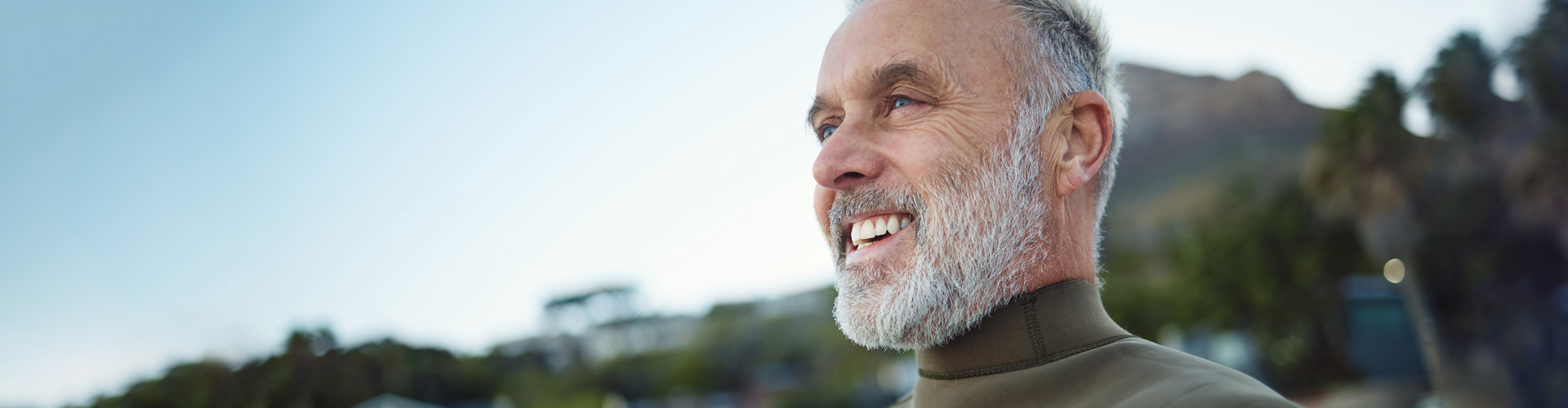 Close-up portrait of a smiling older man with a gray beard outdoors, with trees and hills softly blurred behind him.