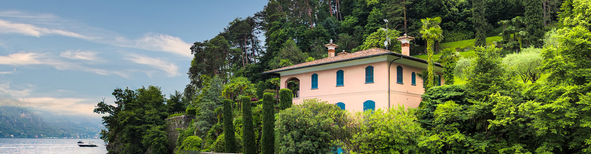 Villa surrounded by lush green trees and gardens on a hillside beside a lake.