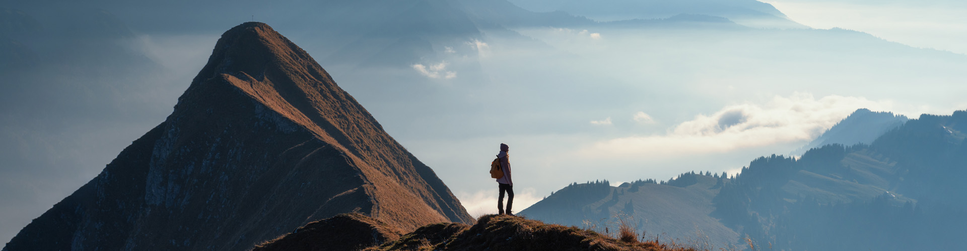 Lone hiker standing on a mountain ridge, looking out over misty blue hills and a steep peak at sunrise or sunset.