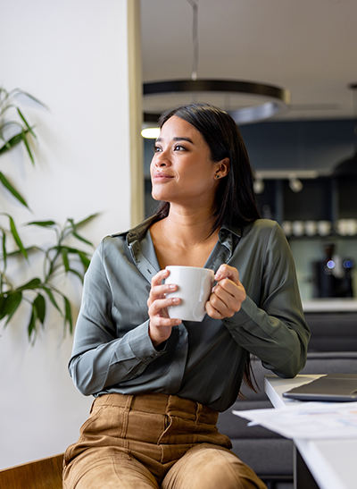 Woman holding a coffee mug at home beside a laptop and paperwork.