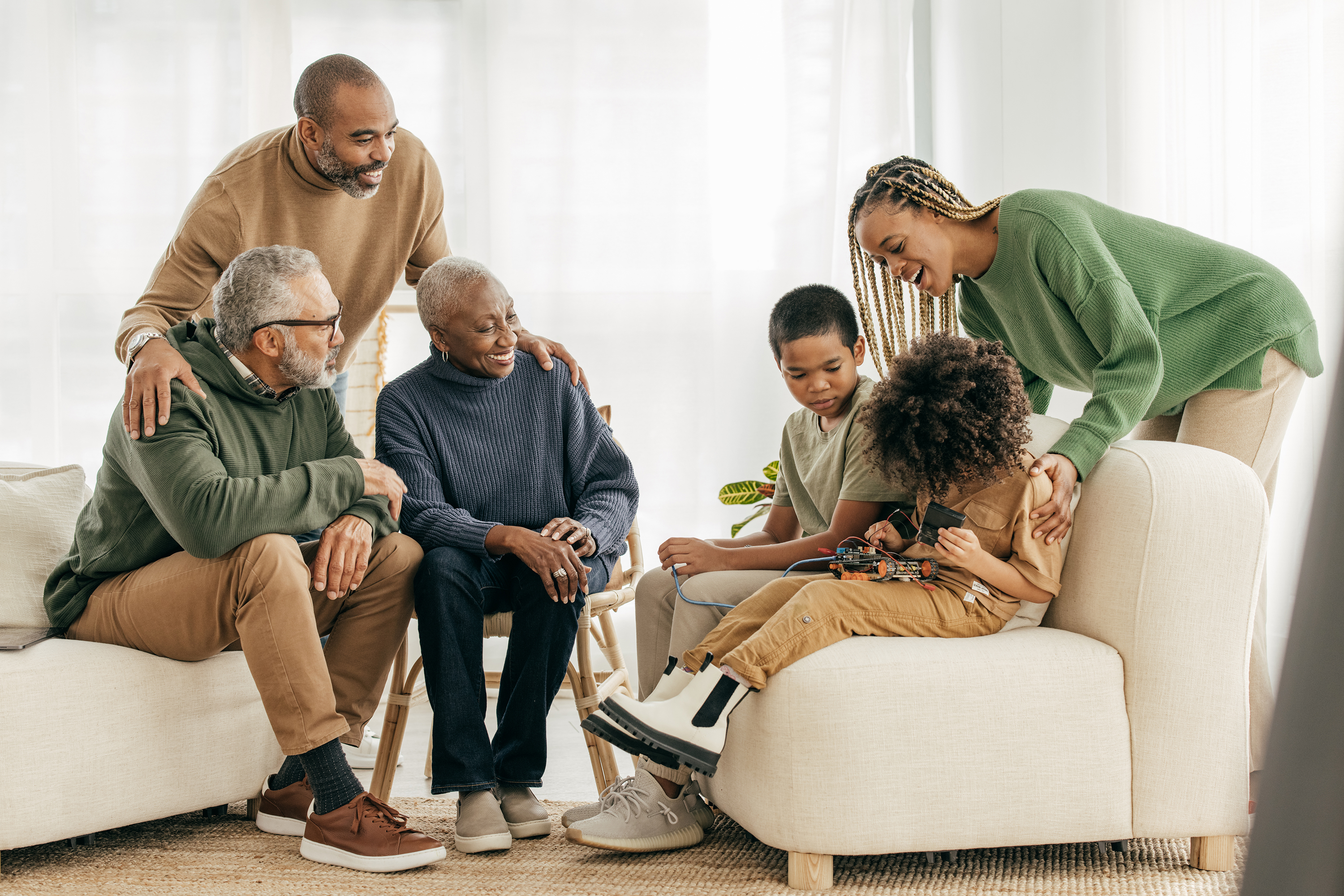 A multigenerational family gathers in a living room while two children work on a small electronics project.