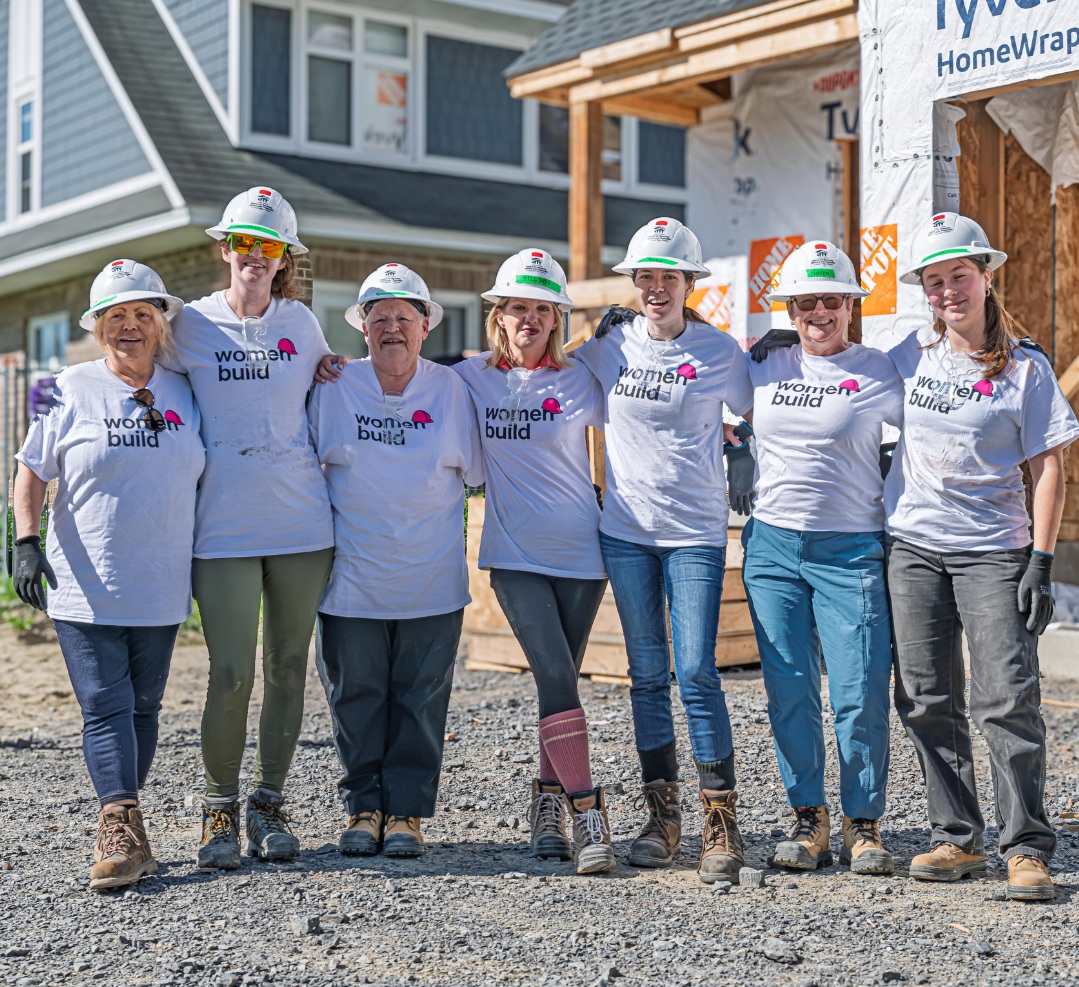 Seven women wearing 'women build' shirts in hard hats on a construction site for Habitat for Humanity build.